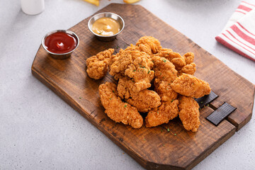 Chicken nuggets or chicken strips on a board with dipping sauces, fries and celery