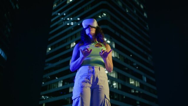 Girl wearing VR glasses moving hands in air, typing on imagine keyboard in cyber space 4K. Low angle shot multi racial young woman working outside at modern company office building night background