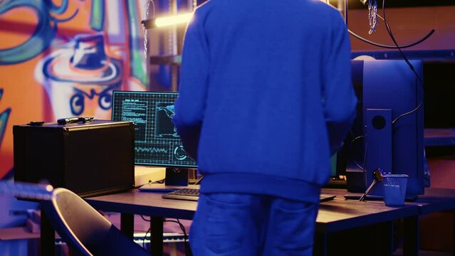 Hacker in abandoned warehouse with graffiti walls writing code that spoofs his location, tricking cops. Rogue developer running script on many devices pinging wrong location to cybercriminal police