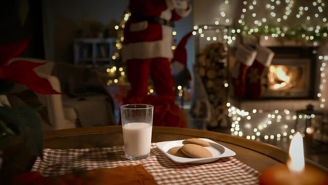 Santa Claus putting Christmas presents in socks over the fireplace, eating ginger cookies and drinking milk on Christmas night in the decorated living room with Christmas tree