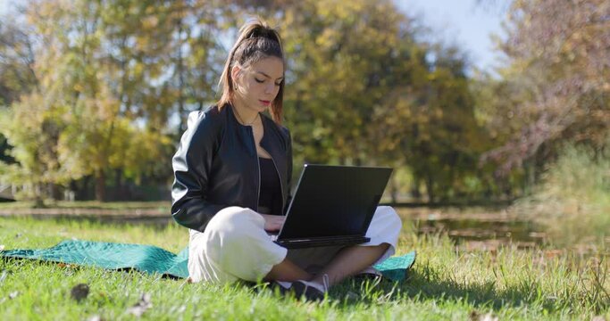 Beautiful girl working on a laptop computer while sitting on a meadow. Fashionable young lady in the park. Left camera movement.