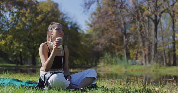 Beautiful girl drinking from a recyclable coffee cup. Fashionable young lady sitting on a field in the park.