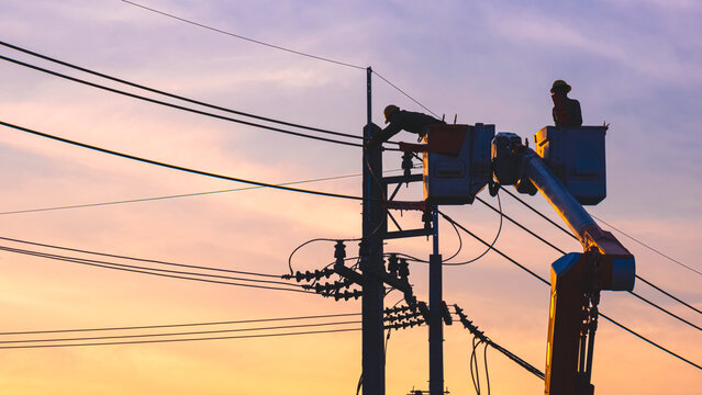 Silhouette of 2 electricians on bucket boom truck are repairing electrical system on electric power pole against sunset sky background in evening time
