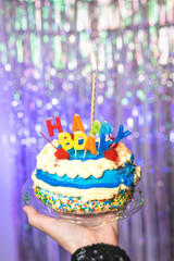 A hand holds up a colorful birthday cake against a unicorn-colored background.