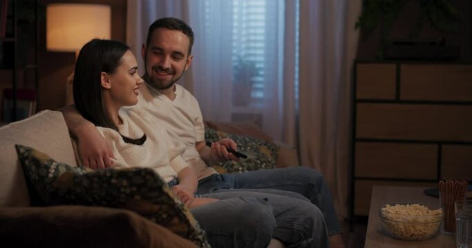 Couple on couch in living room, man with remote control switching TV channels while they eat popcorn. They comment on programs and express boredom or interest.