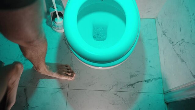 Night, soft blue light in the toilet. A man finishes urinating and flushes the water.