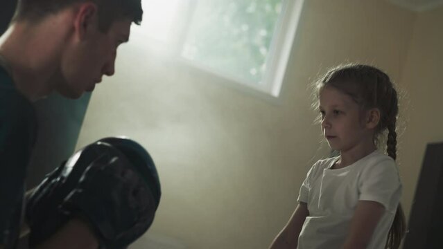 Child punches and blocks with instructor in gym. Young trainer with little girl do fighting exercise in boxing club slow motion. Martial arts school