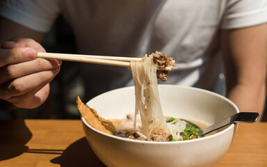 Hand uses chopsticks to pickup tasty rice noodles and pork with noodle in bowl on wooden table. Thai style noodle. Daily light in summer.