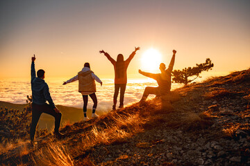 Group of happy friends are standing at mountain top and greeting sunrise or sunset above clouds