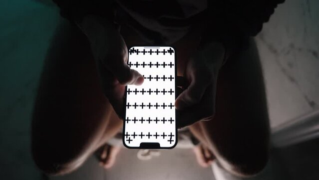 A man in the darkness on the toilet, top view of a smartphone with a white screen for a layout; he uses his fingers to perform a zoom-in gesture on the image. Utilize the white screen for close-up cop