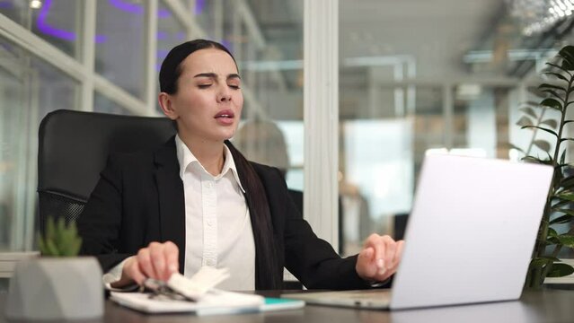 Sick woman freelancer sitting at table near modern laptop and covering mouth with tissue while sneezing indoor. Tired caucasian lady with dark hair working and having runny nose at workplace.