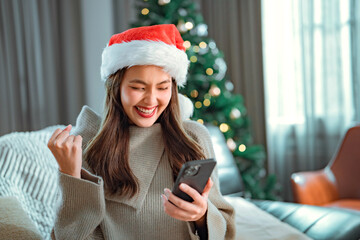 Excited asian woman wearing a Santa hat and using a mobile phone sitting sofa near Christmas tree...