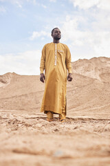 Naklejka premium Dramatic full length portrait of African American man wearing traditional long kaftan in desert landscape