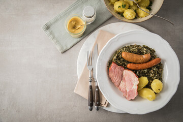 Oldenburger Grünkohl mit geräucherter Kohlwurst, Pinkel, Kassler Nacken, Kartoffeln, Senf, Petersilie auf Teller und hell grau Stein Hintergrund