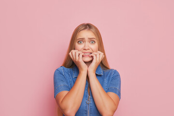 A beautiful girl with long hair poses on pink background in blue jeans top, she is scared and bites...