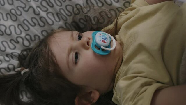 Baby girl sucking a pacifier sitting relaxed on the sofa while holding and playing with a multicolored sheet.