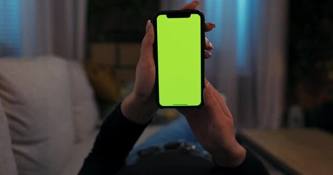 Young woman holding cell phone smartphone in hands observing video photo images while resting on couch sofa surfing Internet watching movie films online .