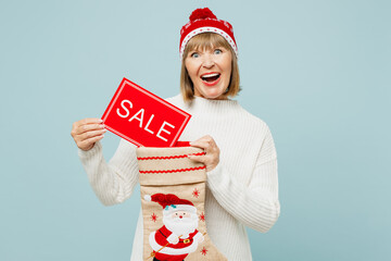 Merry elderly woman 50s years old wear sweater red hat posing hold stocking for gifts, card with sale title text isolated on plain blue background Happy New Year celebration Christmas holiday concept