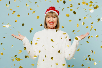 Cheery fun merry elderly woman 50s years old wear sweater red hat posing spread hands stand in confetti rainfall isolated on plain blue background Happy New Year celebration Christmas holiday concept
