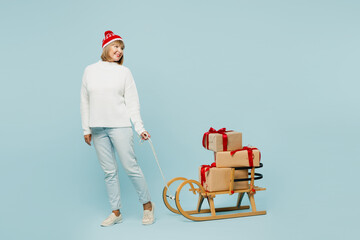 Full body merry elderly woman 50s years old wear sweater red hat posing carry stack of present boxes with gift ribbon bow on sled look aside isolated on plain blue background. Happy New Year concept.