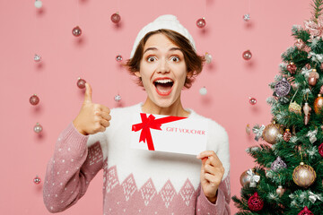Merry young woman in white sweater hat posing hold gift certificate coupon voucher card for store...