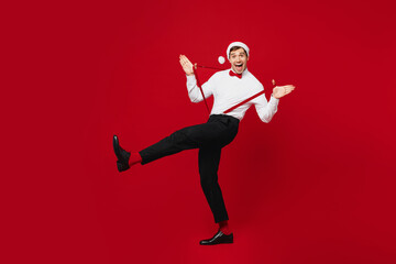 Fototapeta na wymiar Full body side profile view merry young man wear white shirt Santa hat posing hold suspenders raise up leg isolated on plain red background studio Happy New Year Christmas celebration holiday concept
