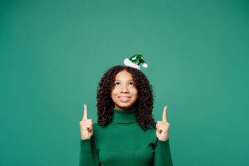 Merry little kid teen girl wears turtleneck hat casual clothes posing point index finger overhead indicate on area copy space isolated on plain green background studio. Happy New Year holiday concept.