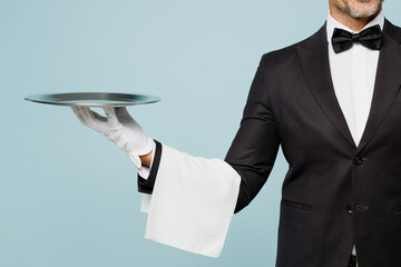 Cropped close up adult barista male waiter butler man wear shirt black suit bow tie uniform hold in hand metal tray plate work at cafe isolated on plain blue background. Restaurant employee concept.