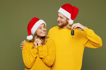 Merry young couple two friends man woman wear sweater Santa hat posing hold in hand car key fob keyless system isolated on plain green background. Happy New Year celebration Christmas holiday concept.