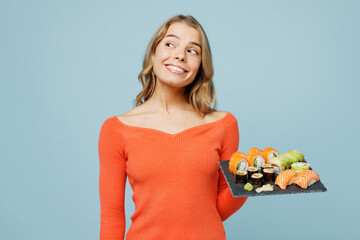 Young smiling impressed woman wearing orange casual clothes ook aside on workspace area hold eat fresh raw makizushi sushi roll served on black plate Japanese food isolated on plain blue background.