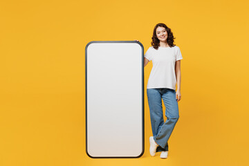 Full body young smiling fun woman she wear white blank t-shirt casual clothes stand near big huge blank screen mobile cell phone smartphone with area isolated on plain yellow orange background studio.