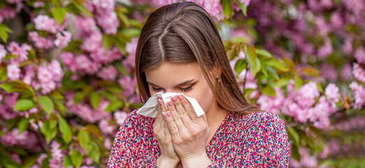 Spring allergy concept. Sneezing girl with nose wiper among blooming trees in park. Pollen allergy,...