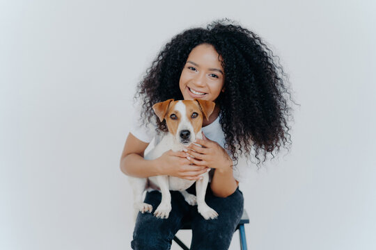 Cheerful young Black woman with curly hair hugging her Jack Russell Terrier, both looking happy against a white background