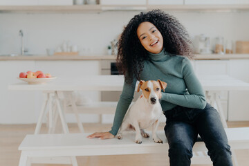 Smiling woman in turtleneck with her dog at a kitchen table, radiating joy.