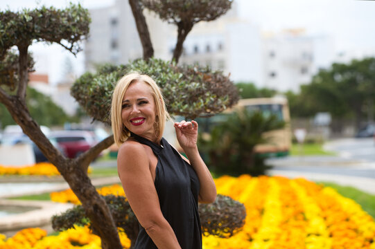 Mature, blonde and beautiful woman wears an elegant black dress and poses for photos next to a garden with an olive tree and flowers. The woman is happy and smiling while doing different expressions.