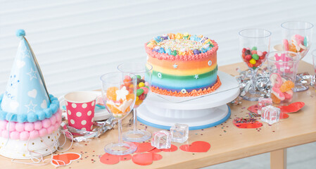 Cake on birthday with rainbow butter cream decorated with sugar candies, colorful sprinkles. Beautiful cake placed on stand with colorful candies, jelly in glasses and hats on table. Festive celebrate