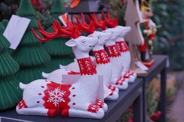  a row of decorative figures of white deer with red ornaments next to green Christmas trees on a shelf in a store before Christmas