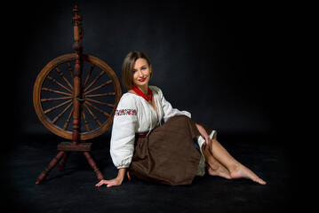 Fototapeta na wymiar beautiful ukrainian brunette smiling woman in ukrainian vintage traditional dress embroidery and red necklace sitting barefoot near retro spinning wheel and holding it In her hand on black background 