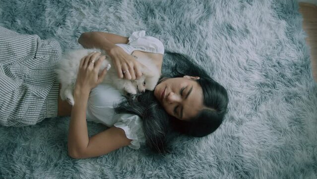 Young thai woman holding cute little white spitz dog lying on the carpet in living room with modern interior