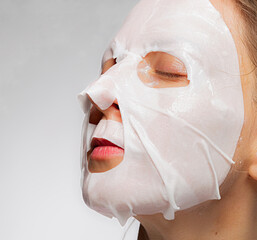 Woman with a sheet moisturizing mask on her face. Woman applying cotton facial moisturizing mask on face, takes care of skin, prevents wrinkles, stands on isolated gray background.