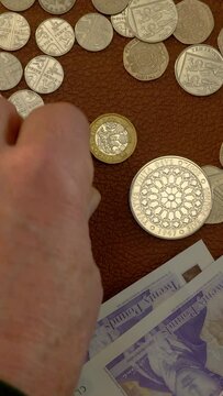 Vertical video social media format – Closeup overhead shot of a rare £5 silver coin spinning and falling among other sterling currency, then a man’s hand adding some pound coins.