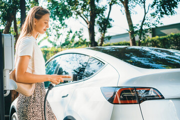 Young woman travel with EV electric car charging in green sustainable city outdoor garden in...