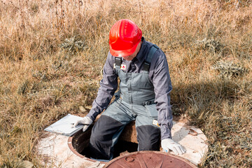 A plumber in overalls and a helmet sits on the side of a well hatch with documents, checking the...