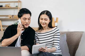 Asian couple relaxing with smartphone at home, loving man and woman sitting on couch together, Browsing Internet on mobile phone and laptop.