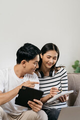 Asian couple relaxing with smartphone at home, loving man and woman sitting on couch together, Browsing Internet on mobile phone and laptop.