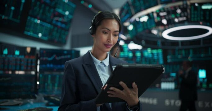 Portrait of a Beautiful Asian Female Working in an International Stock Exchange Hall: Specialist Wearing Headphones and Using Tablet Computer, Successful Trader Looking at Camera and Smiling