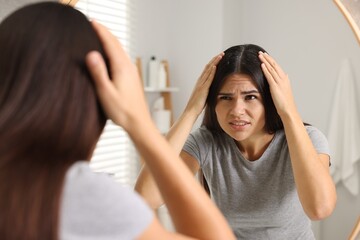 Emotional woman examining her hair and scalp near mirror in bathroom. Dandruff problem