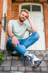 Smiling and handsome young male.Handsome young man looking at camera with smile while sitting on the stairs near brick wall house.Summer hot day.