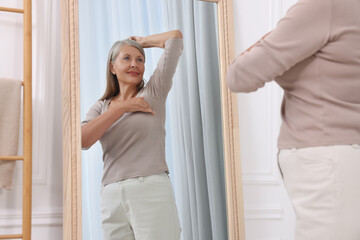 Fototapeta na wymiar Beautiful senior woman doing breast self-examination near mirror indoors
