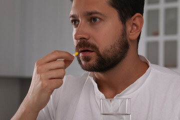 Depressed man with glass of water taking antidepressant pill indoors, closeup
